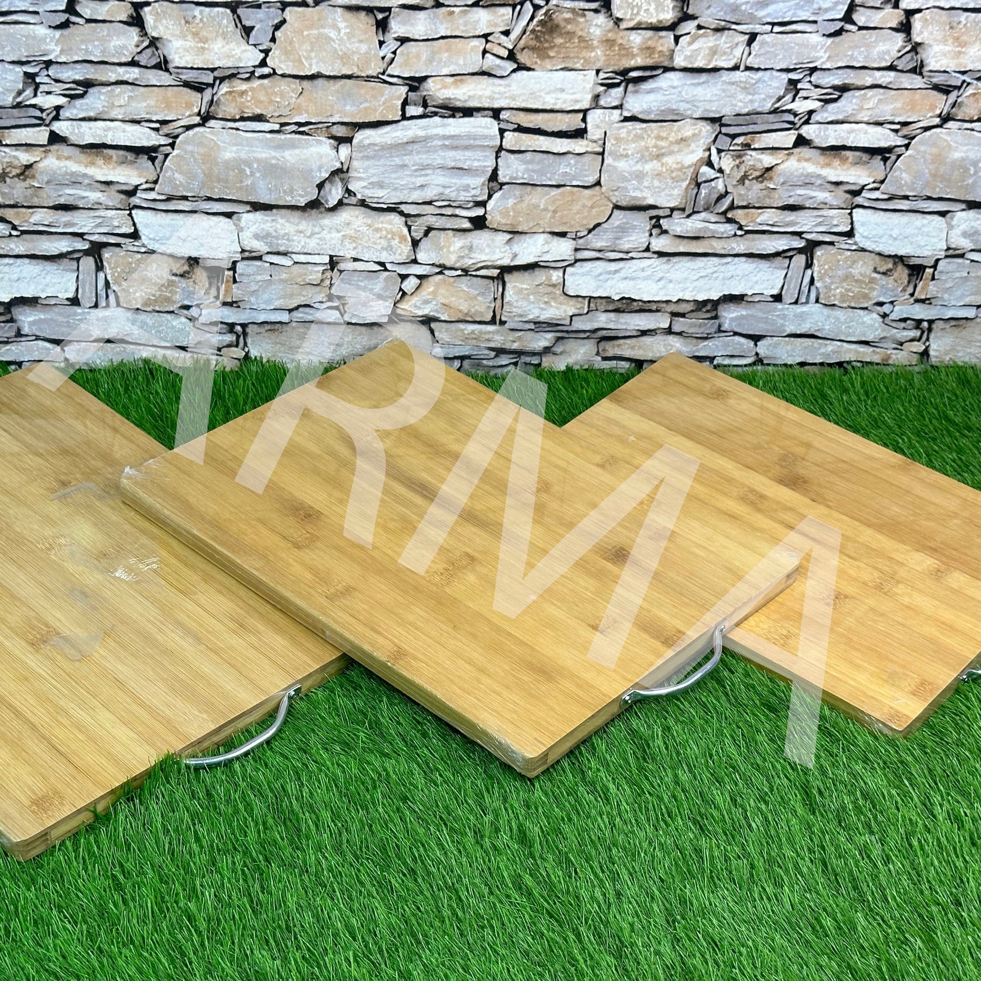 Three square-shaped bamboo cutting boards of different sizes, displayed on a grassy surface against a stone wall.