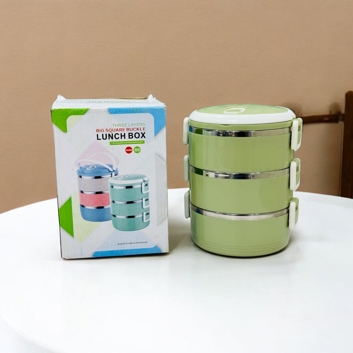 A 3-layer stainless steel lunch box in green color, displayed alongside its packaging which has 'Lunch Box' text and images showing the product in use.